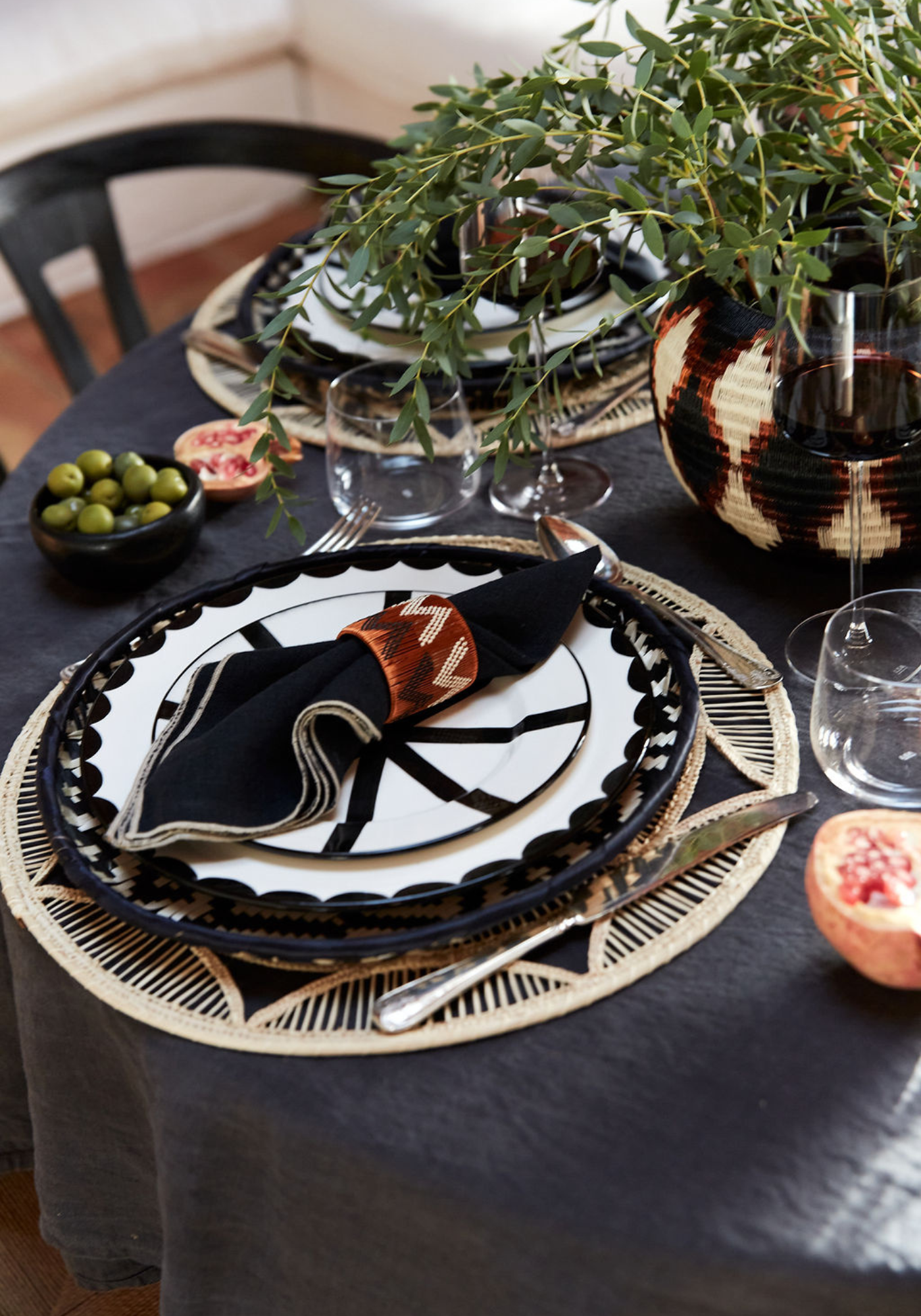Scandi Festivities tablescape featuring black and ivory geometric hand-painted plates, woven rattan placemats, black linen napkins with terracotta rings, and greenery in a woven centrepiece.