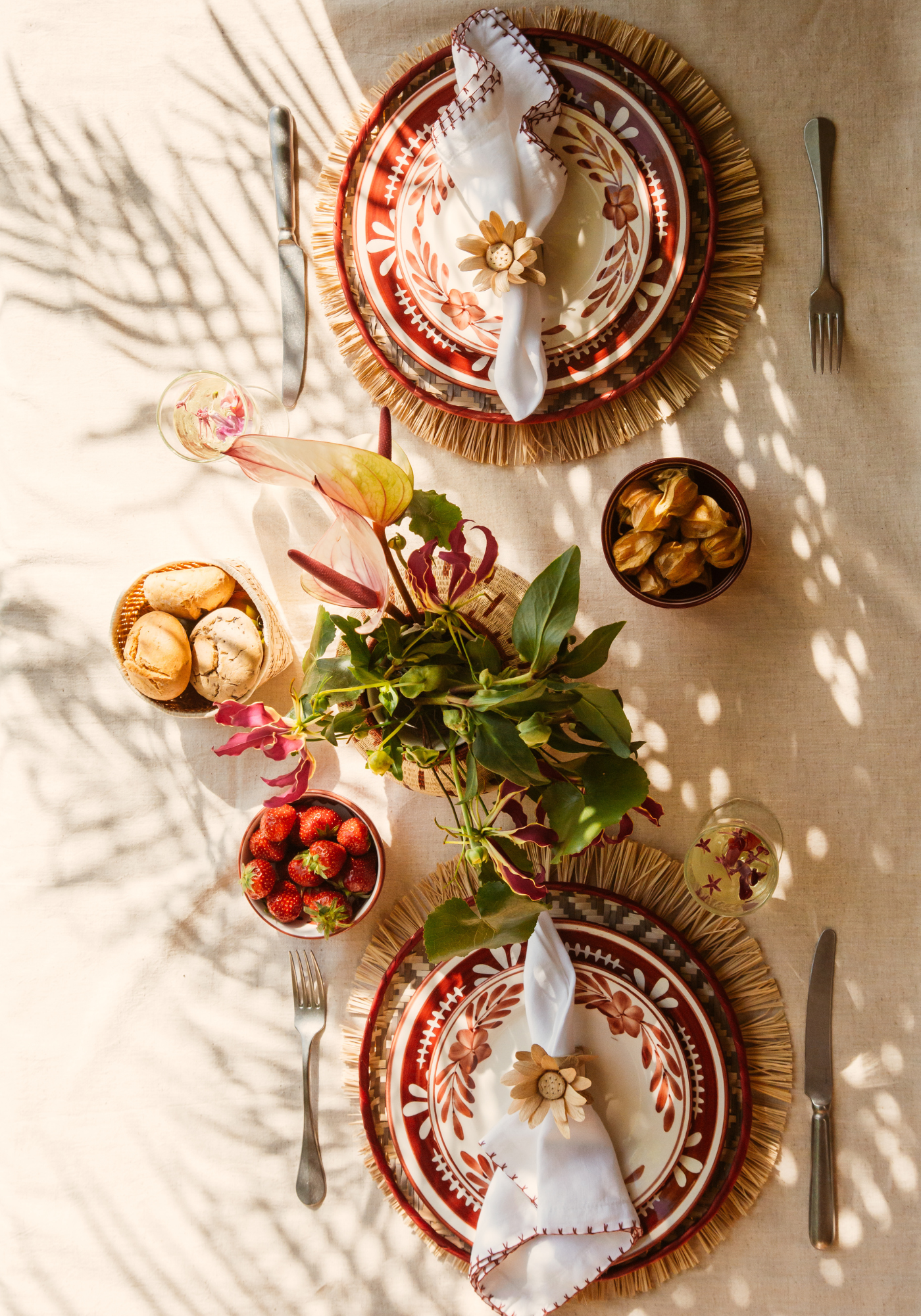 Bohemian Brunch tablescape with terracotta hand-painted floral plates, straw-edged woven placemats, white napkins with red stitching, and flower napkin rings surrounded by tropical fruit and flowers.