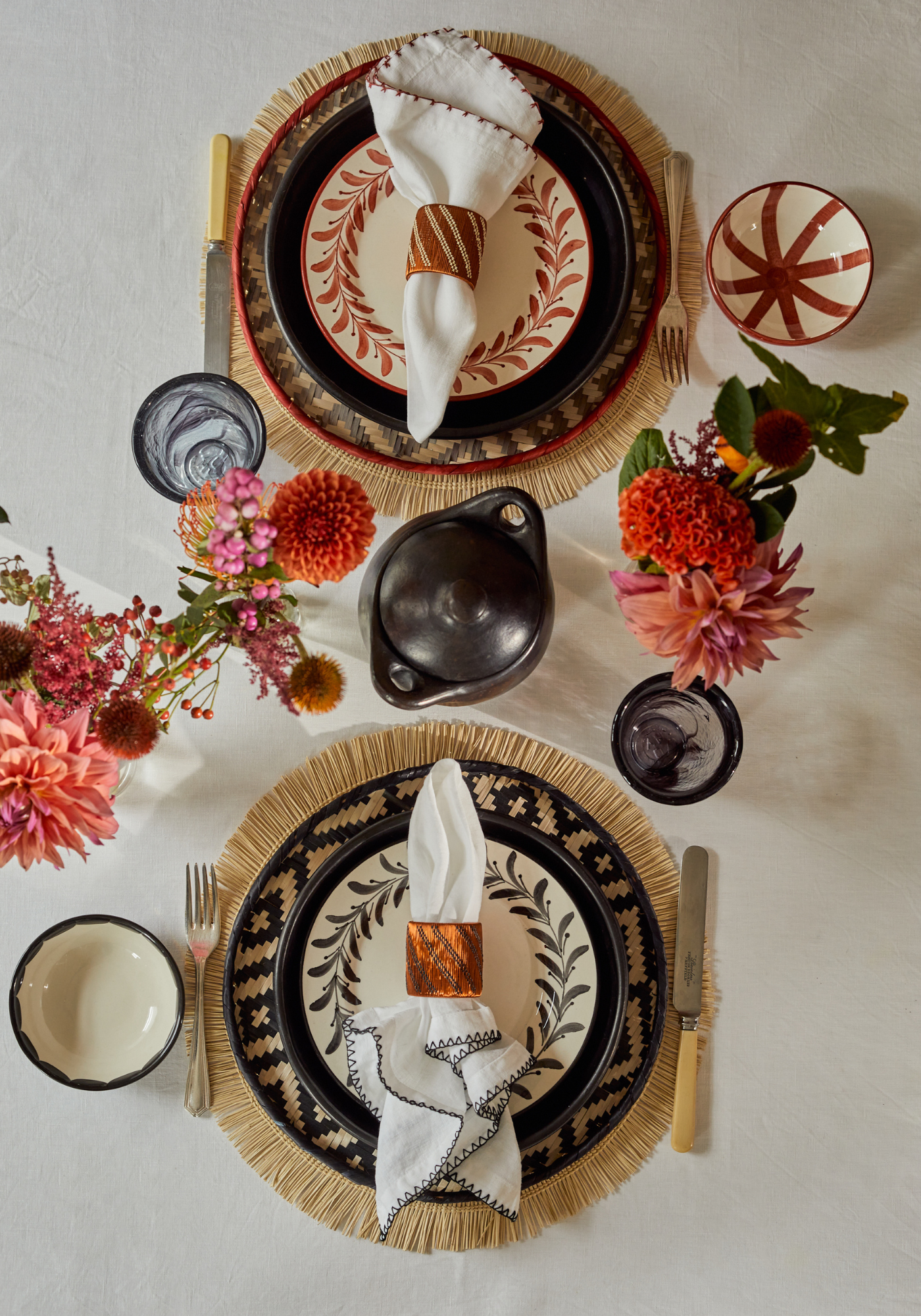Sunday Best tablescape featuring terracotta and black hand-painted ceramics, straw-edged woven placemats, contrasting napkins with embroidered edges, and bright dahlias on a white linen tablecloth.