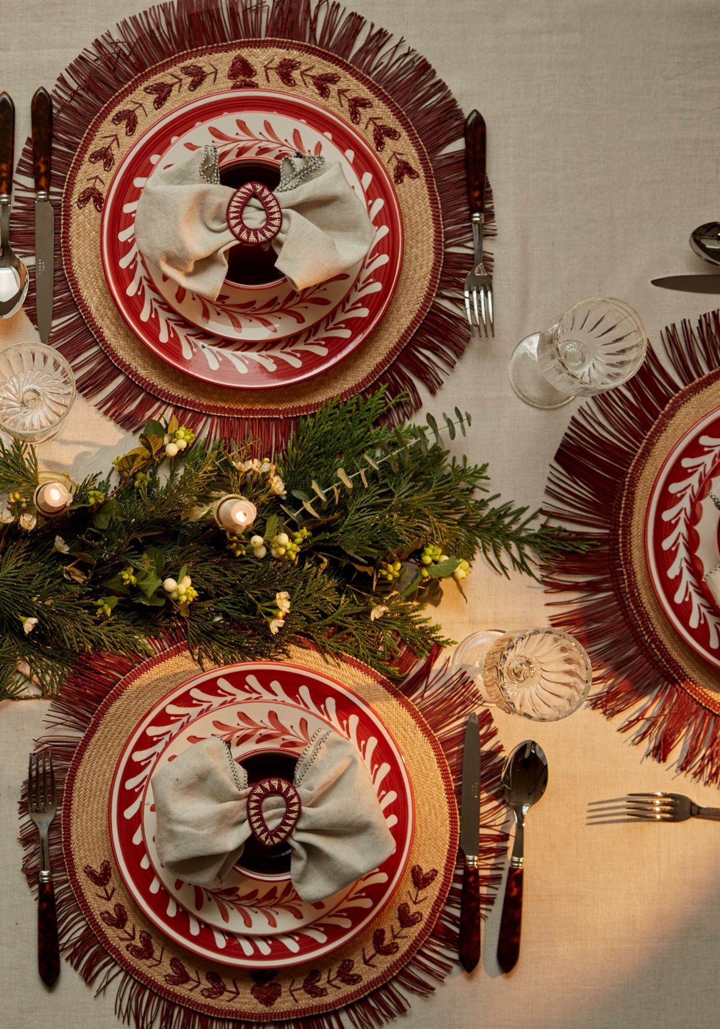 Nordic Christmas tablescape with embroidered marsala placemats, rust-red Helecho plates, linen napkins with marsala Gota rings, crystal glassware, and an evergreen candlelit centrepiece.