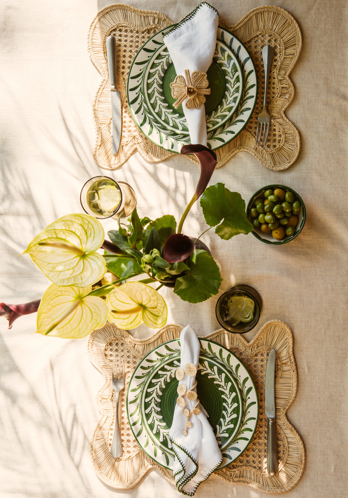 Lunch au Natural tablescape featuring hand-painted green ceramic plates, sculptural rattan placemats, white linen napkins with green stitching, and natural fibre napkin rings with calla lilies.