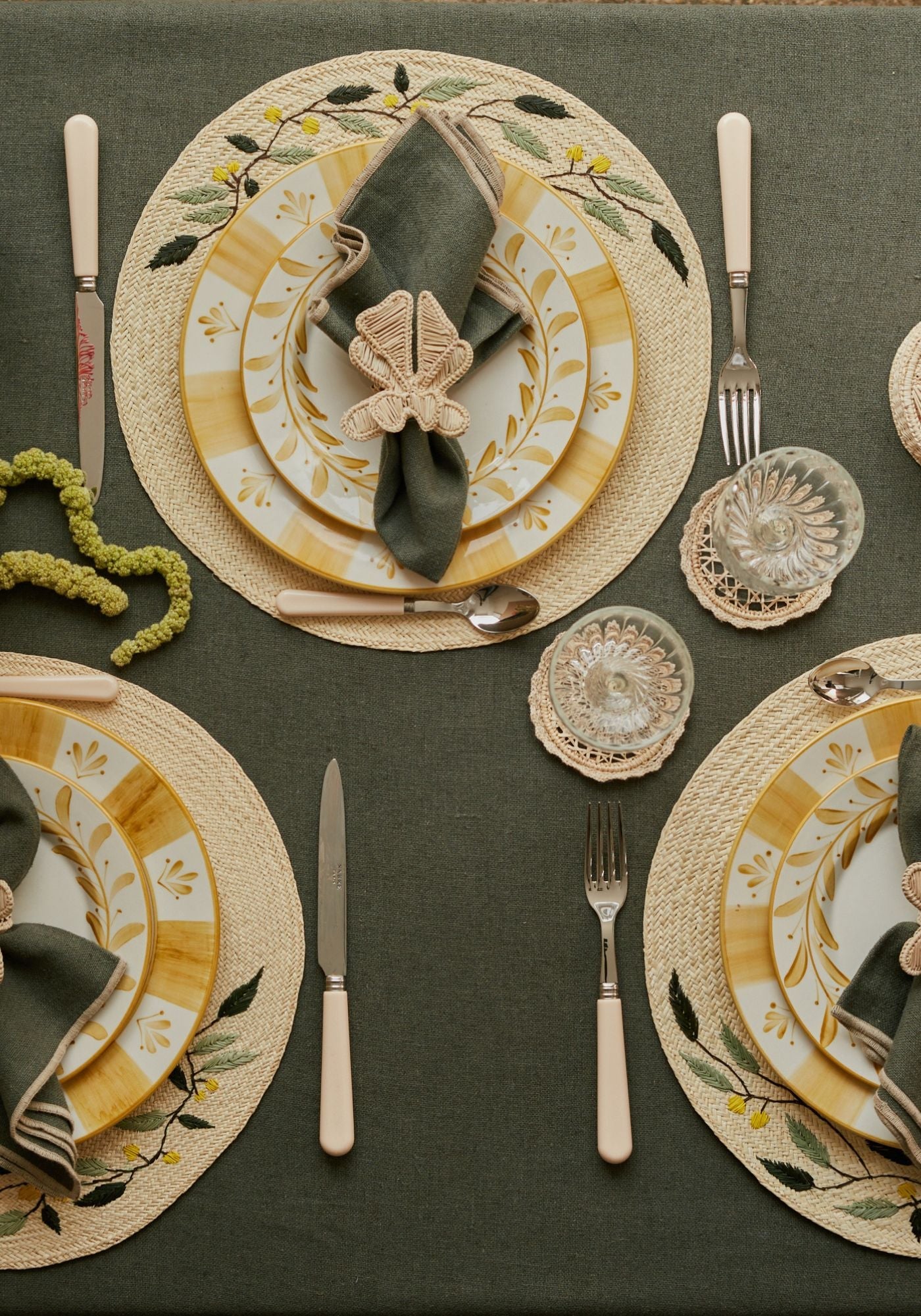 Festive tablescape featuring embroidered Leaf placemats, golden yellow Carmen plates, green linen napkins with Ribbon napkin rings, clear glassware, and cream-handled cutlery styled on a deep green tablecloth.