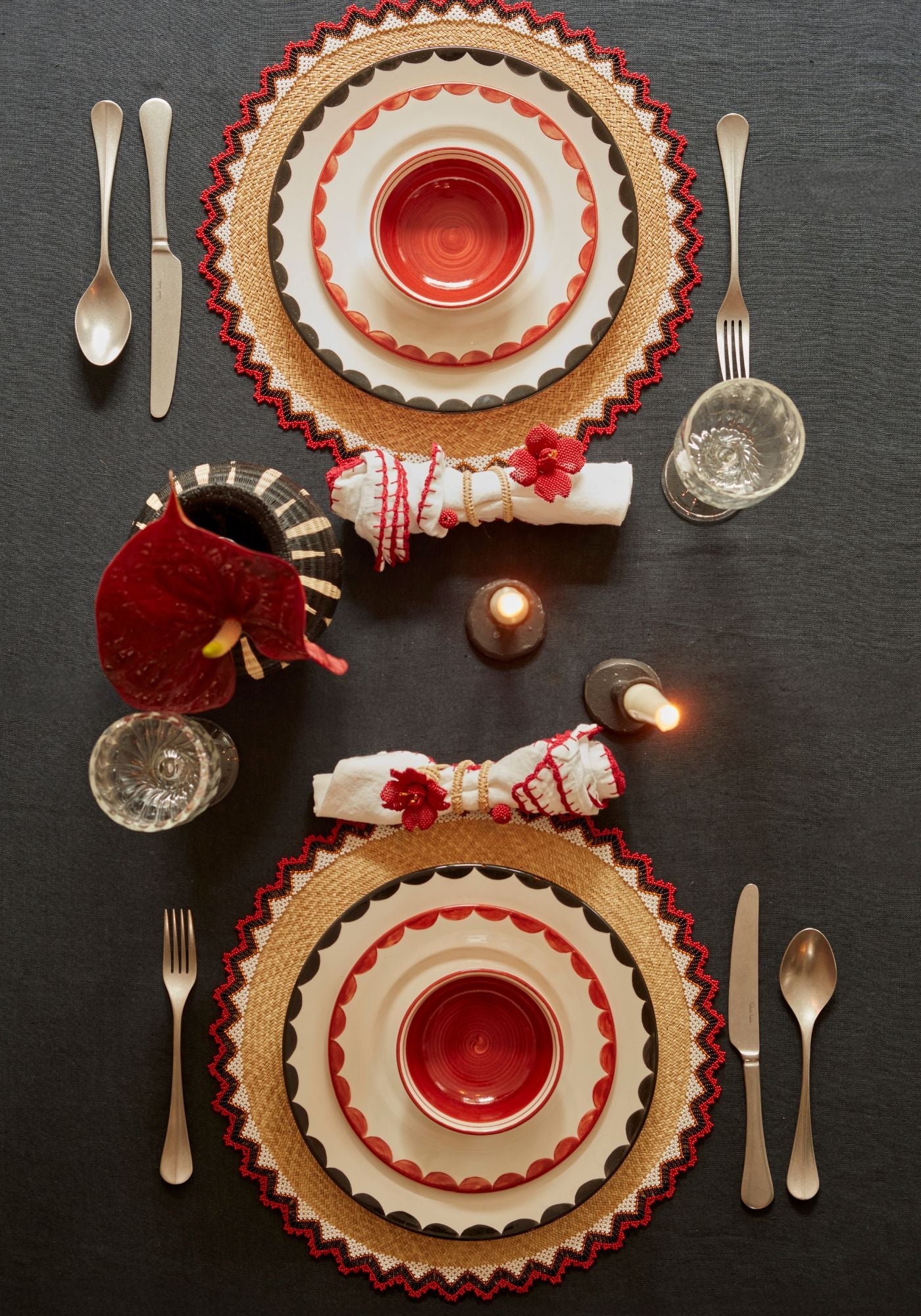 Top view of a refined festive tablescape featuring CasaLatina’s handmade red and black ceramic plates layered on woven placemats with beaded trims, white napkins adorned with red accents, candles, and crystal glassware on a black linen tablecloth.