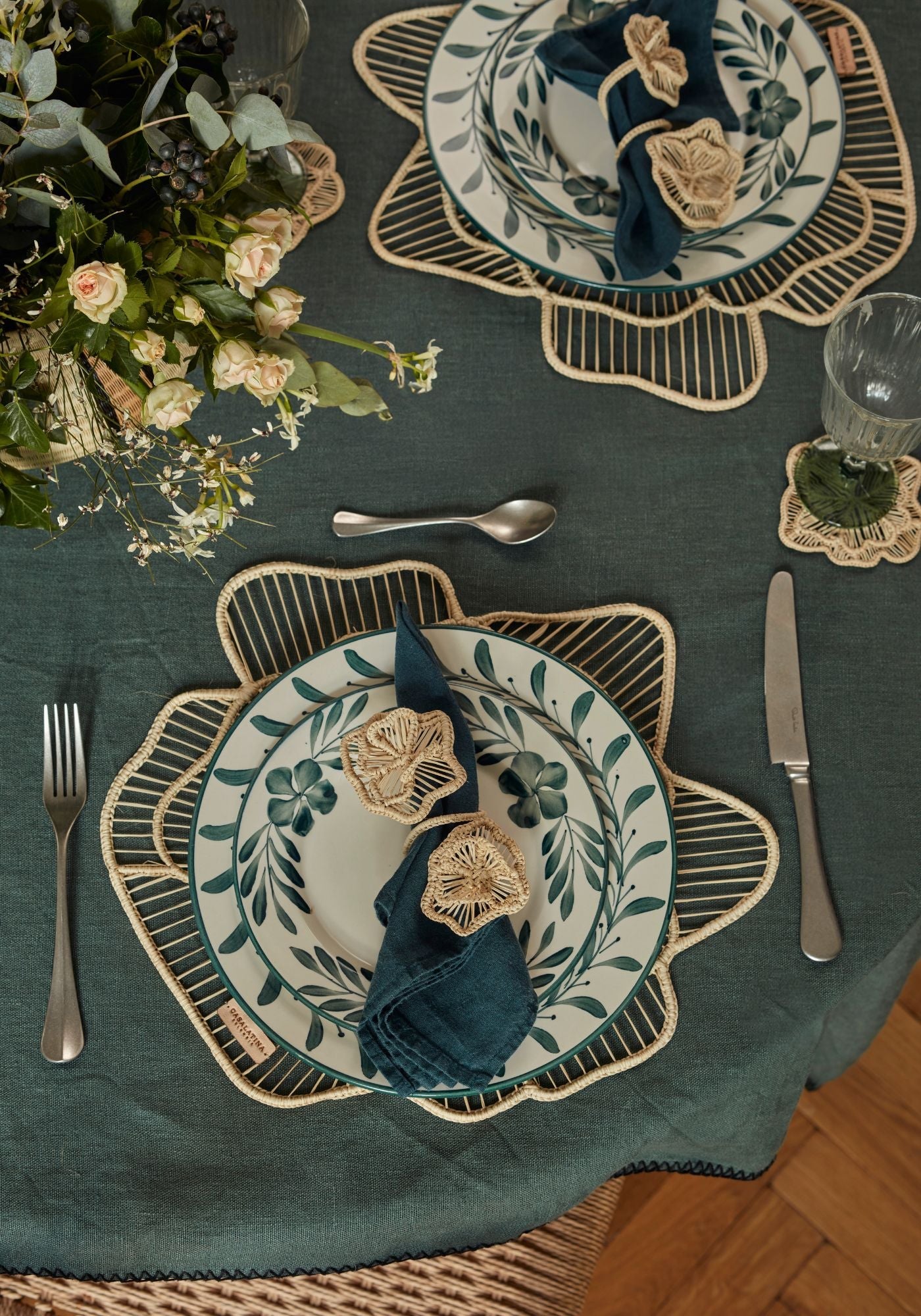Say It With Flowers tablescape featuring teal floral hand-painted plates, petal-shaped woven placemats, and raffia flower napkin rings on a dark teal linen tablecloth.
