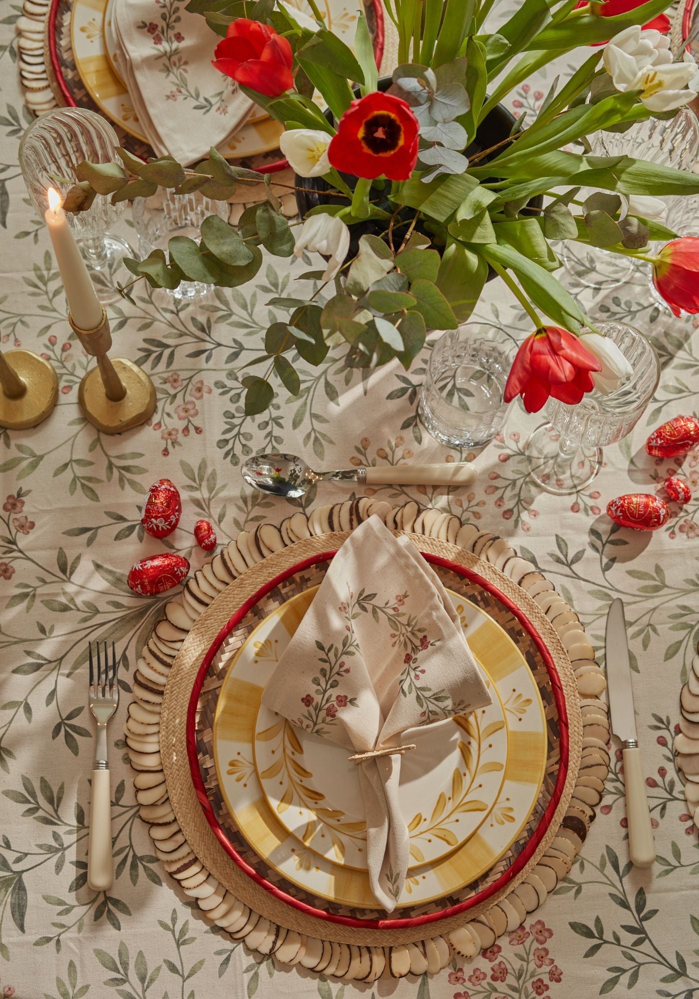 Easter Lunch tablescape with floral linen tablecloth, yellow hand-painted plates, woven placemats with tagua trim, and bunny-ear napkin rings styled with tulips and candles.