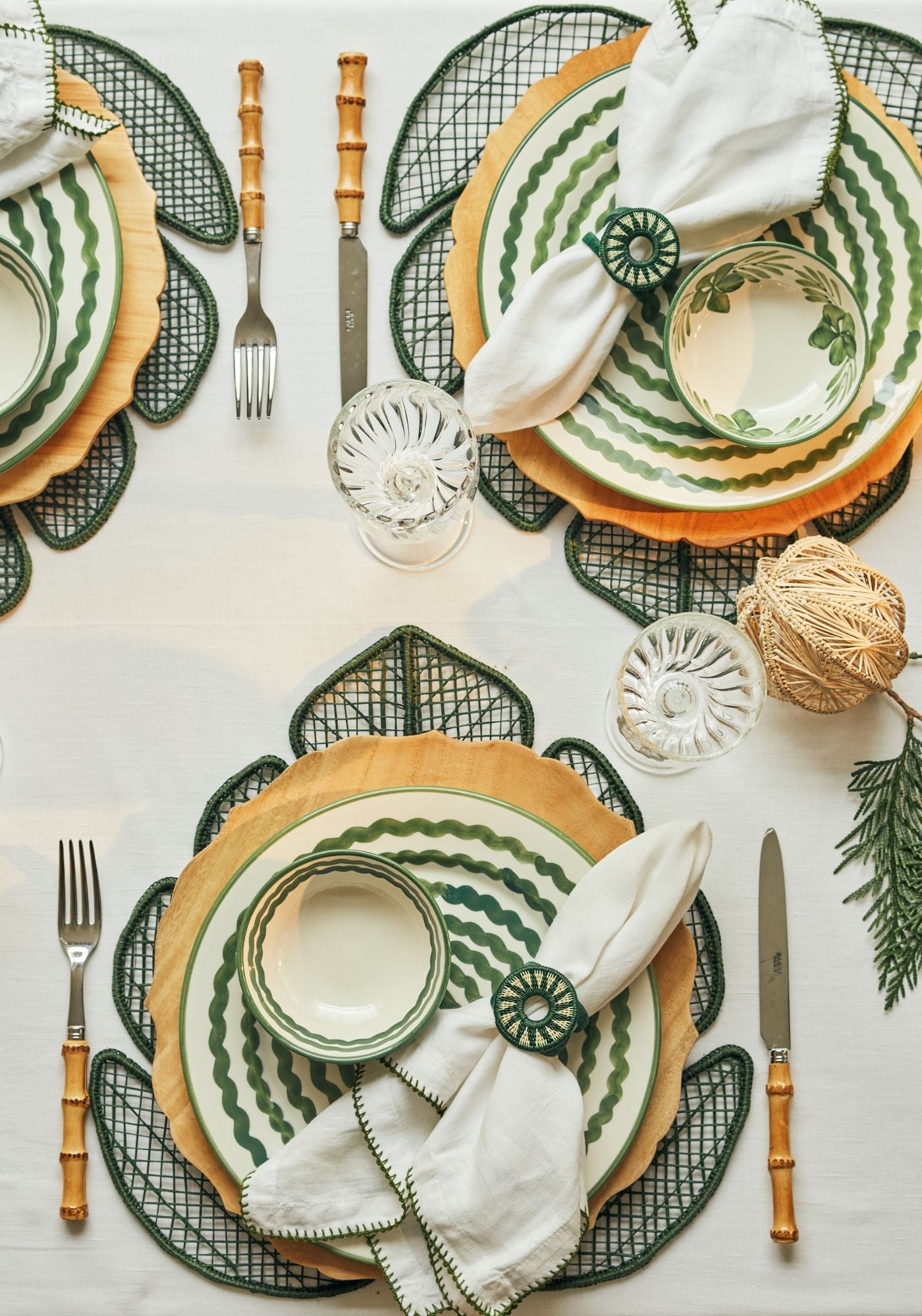 Alpine breakfast tablescape with green Hoja placemats, wooden Selva bevelled chargers, Magdalena hand-painted plates in Cascos and Ondas designs, white linen napkins with green Sol napkin rings, and clear glassware on a white tablecloth.